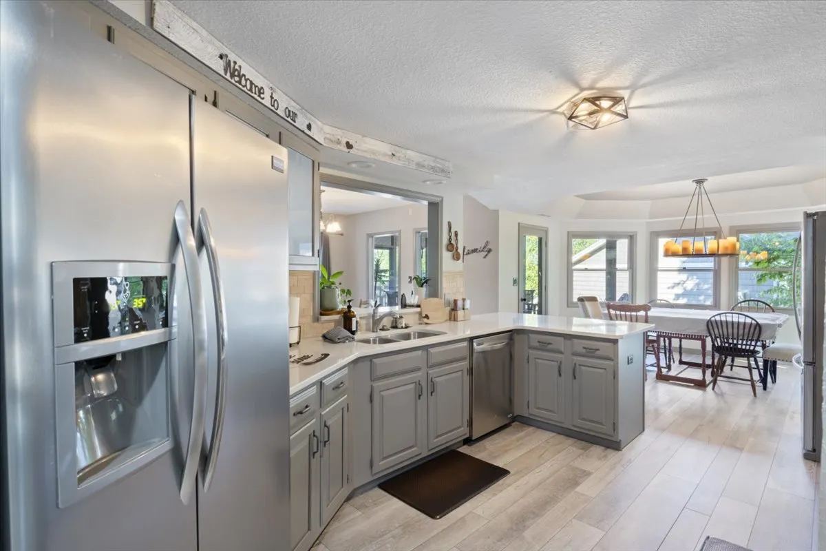 A bright kitchen illuminated by natural light, featuring a stainless steel refrigerator, expansive white countertops, and a dining area visible in the background. The kitchen is neatly arranged and clearly designed for both cooking and entertaining, with modern finishes and open space for meal preparation.