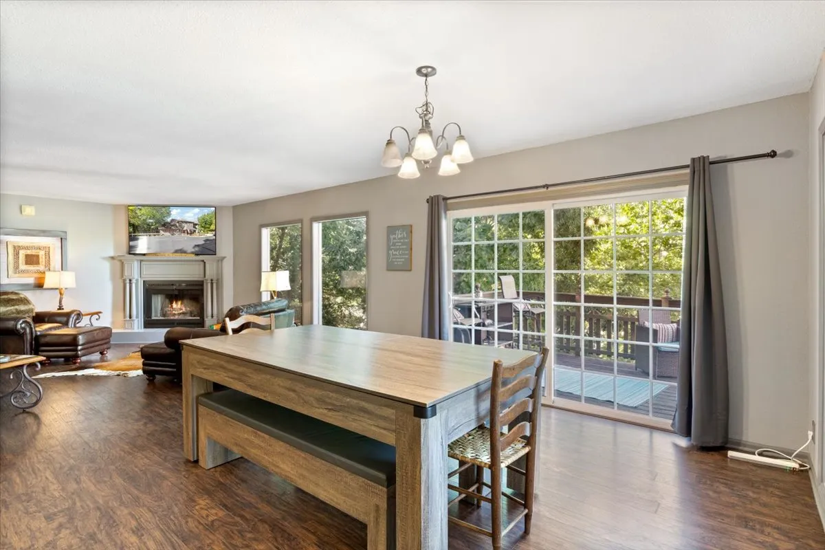 A bright, comfortable dining area anchored by a substantial table, set near a fireplace, with large windows looking out onto an outdoor deck. The space feels inviting and ideal for both shared meals and relaxing moments.