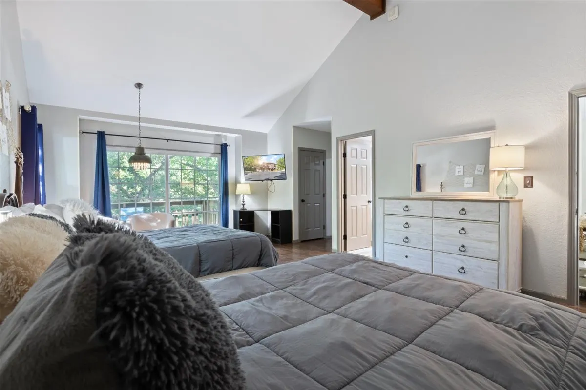 A tranquil bedroom with a large window streaming natural light onto a comfortable bed with plush bedding. The room also features a decorative pendant light, a dresser with a mirror, and subtle decorative accents, all creating an inviting space for relaxation.