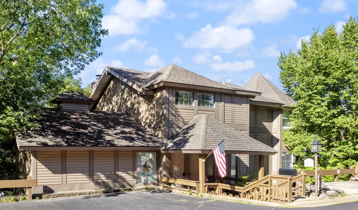 The hero image showcases the welcoming front entrance of a spacious, beautifully landscaped single-family lodge. An American flag waves beside inviting architectural features, while lush gardens and a wide porch hint at spaces perfect for relaxing and socializing. The warm, well-maintained exterior sets a friendly and impressive first impression.