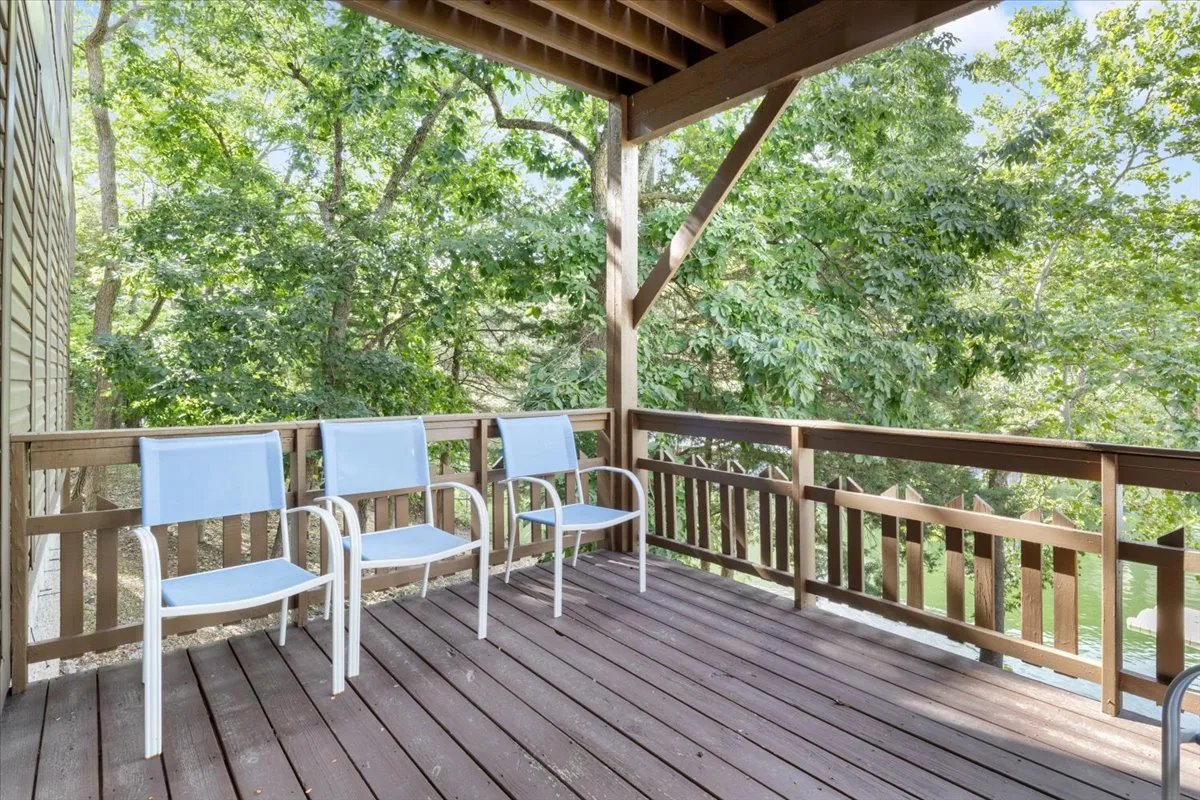 A peaceful wooden deck shaded by trees, furnished with white chairs and surrounded by lush greenery. The space overlooks natural scenery, suggesting a perfect spot for groups to gather, relax, and share stories while enjoying the outdoors. Hints of dawn or dusk lighting evoke a sense of togetherness and tranquility.